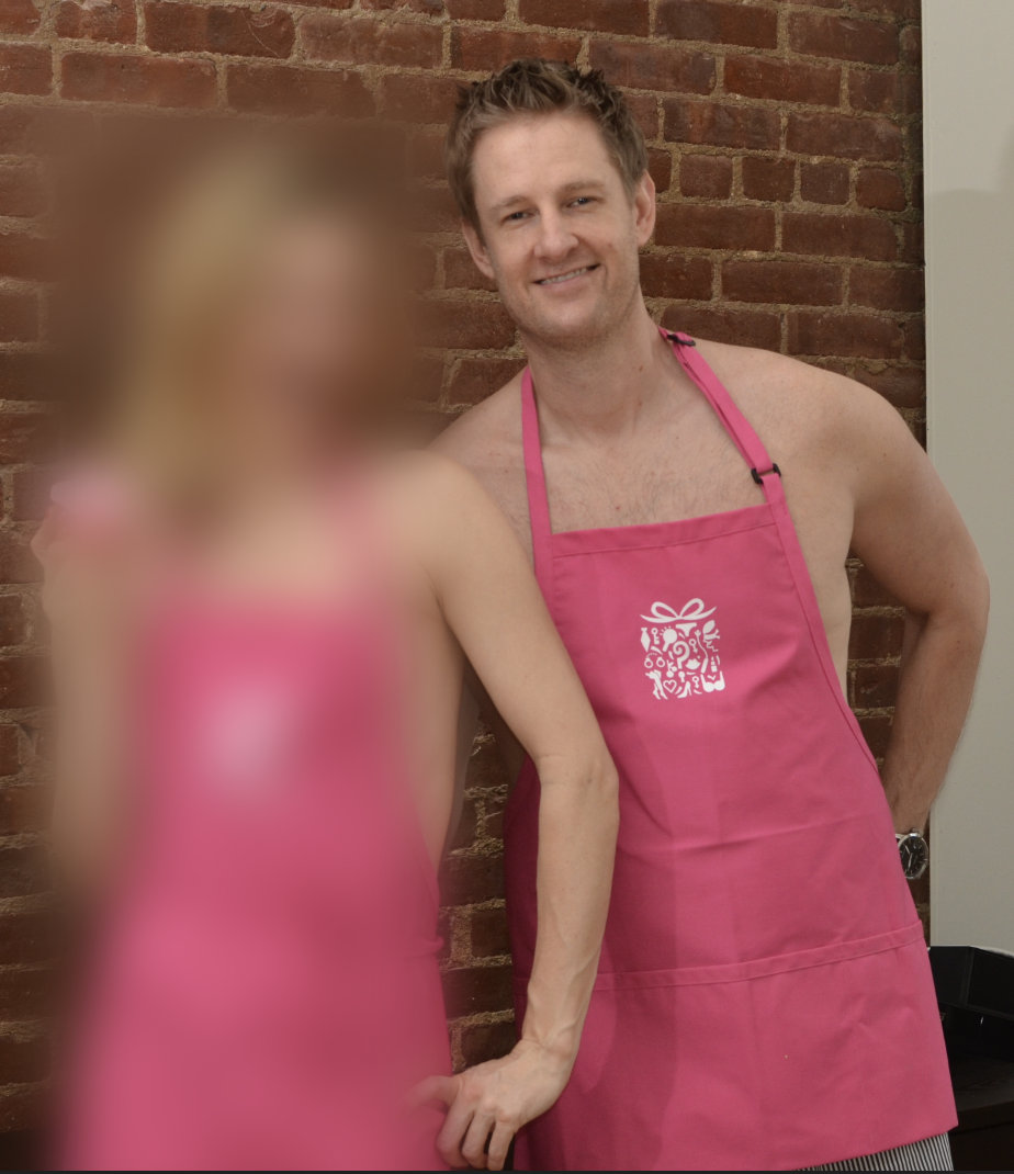 A man with short brown hair smiling, wearing a pink apron with a white gift box logo, standing next to a woman with blonde hair in a pink apron, against a brick wall background.