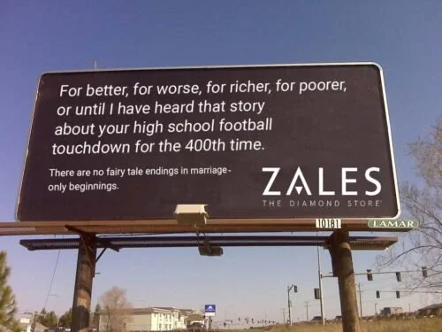 Large billboard with Zales Diamond Store logo, displaying a humorous message about high school football touchdowns and fairy tale endings, with a clear blue sky and trees in the background.