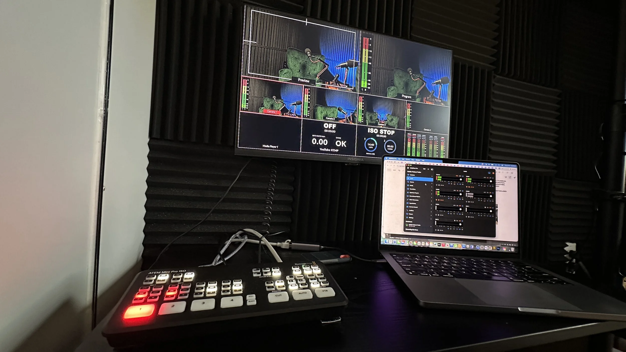 A video production monitor displaying multiple camera angles of a green armchair with a microphone, set against a soundproofed wall with black foam panels. A laptop and a control panel with illuminated buttons are on the desk below the monitor.