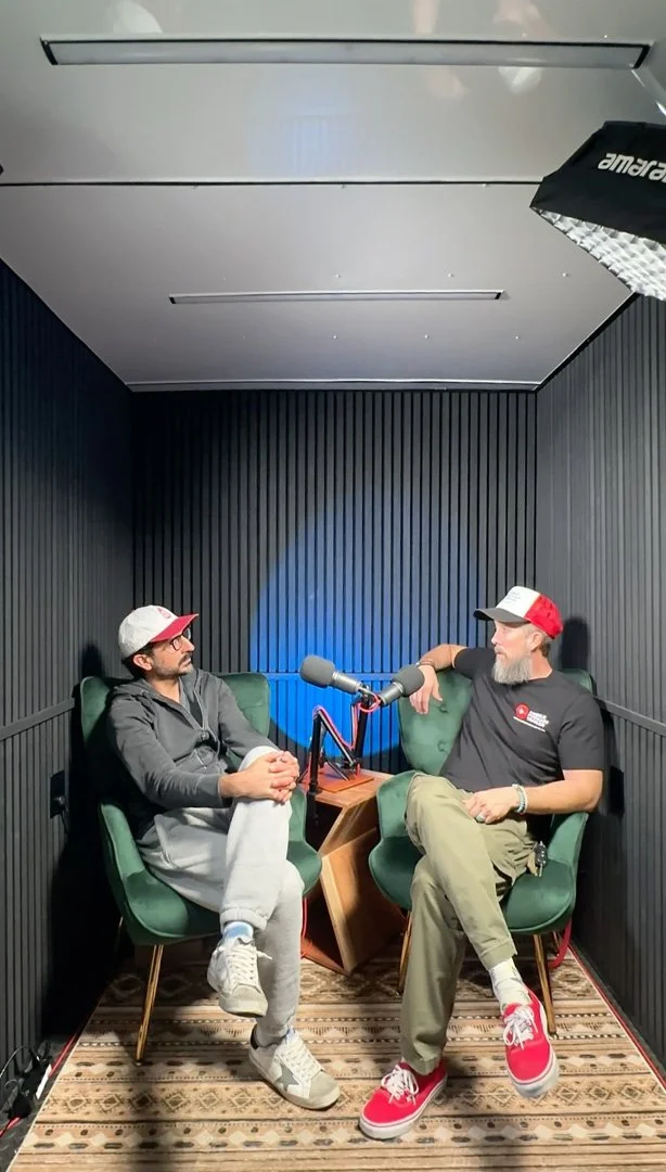 Ivan and Jesse sitting in the Mobile Podcast Trailer with black acoustic panels, having a conversation with a microphone between them.