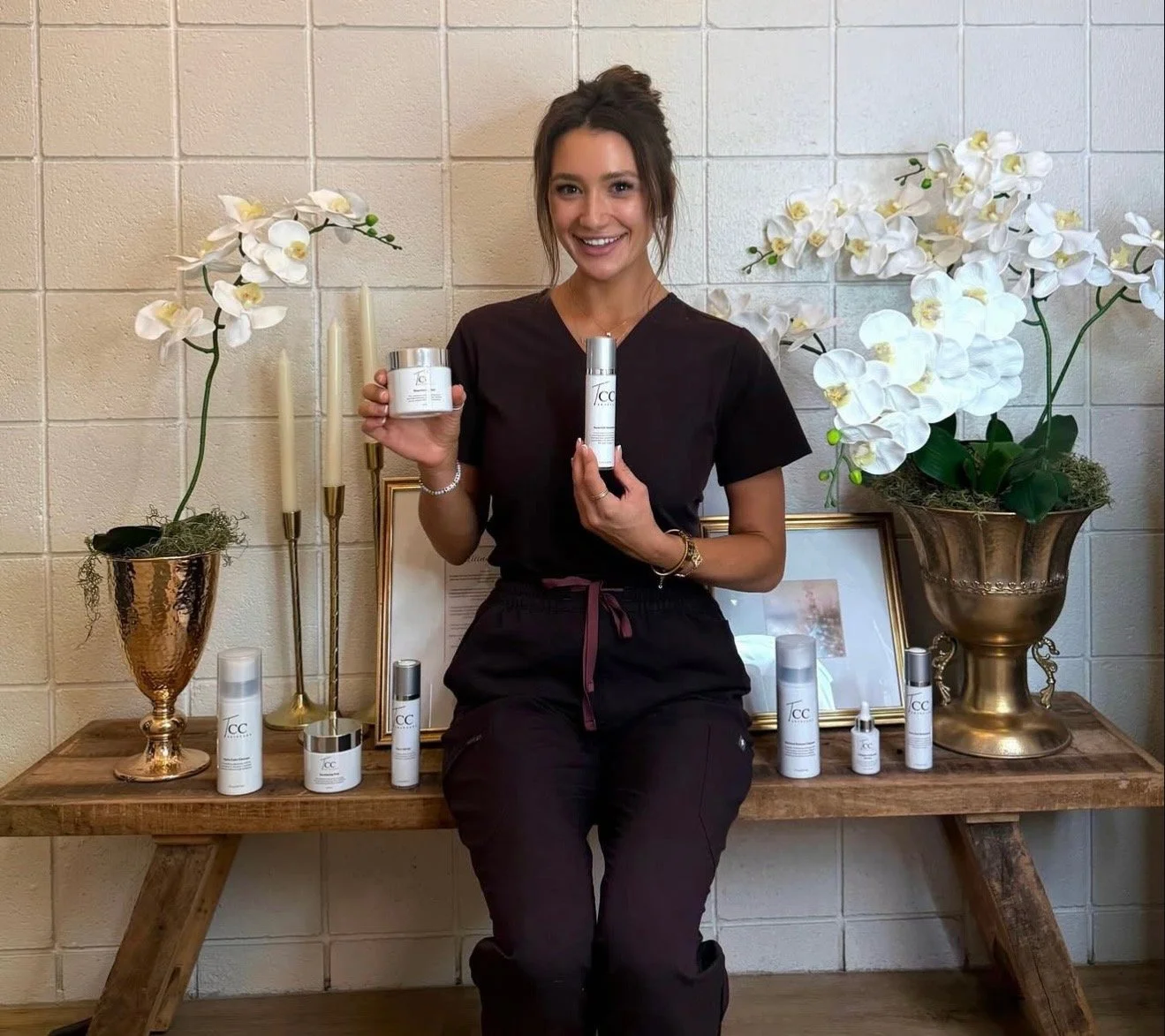 A woman dressed in black scrubs sitting on a wooden bench holding skincare products, with white orchids and candles on either side, and a framed certificate or artwork in the background.