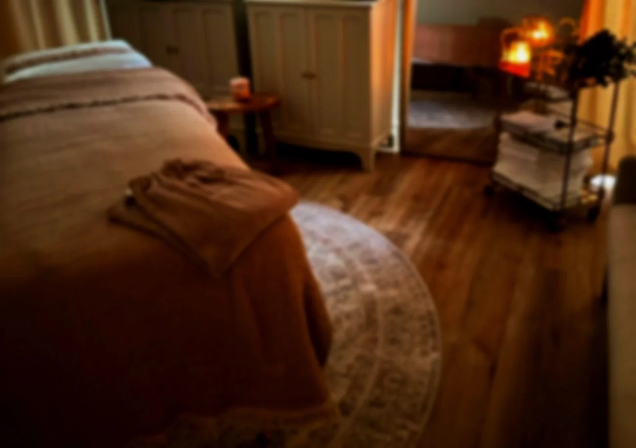 Cozy bedroom with wooden floors, a large bed with a striped blanket, a nightstand with a small lamp, a rounded rug, a wooden wardrobe, and a sporting goods cart with magazines or books in a warm, dimly lit setting.