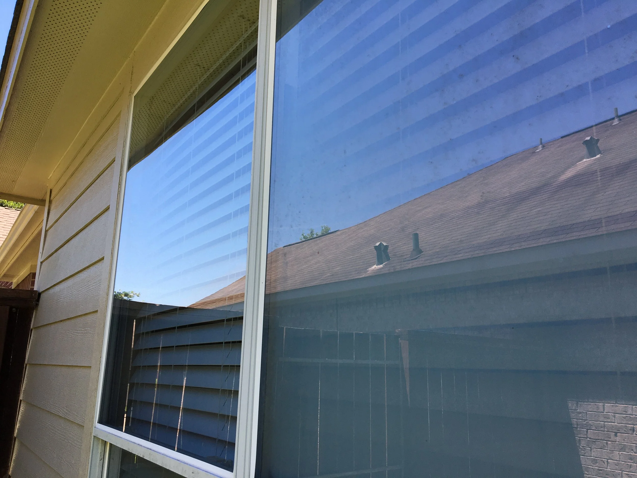 The exterior of a house with beige siding and a large window that reflects the sky, neighboring house roof, and some trees.