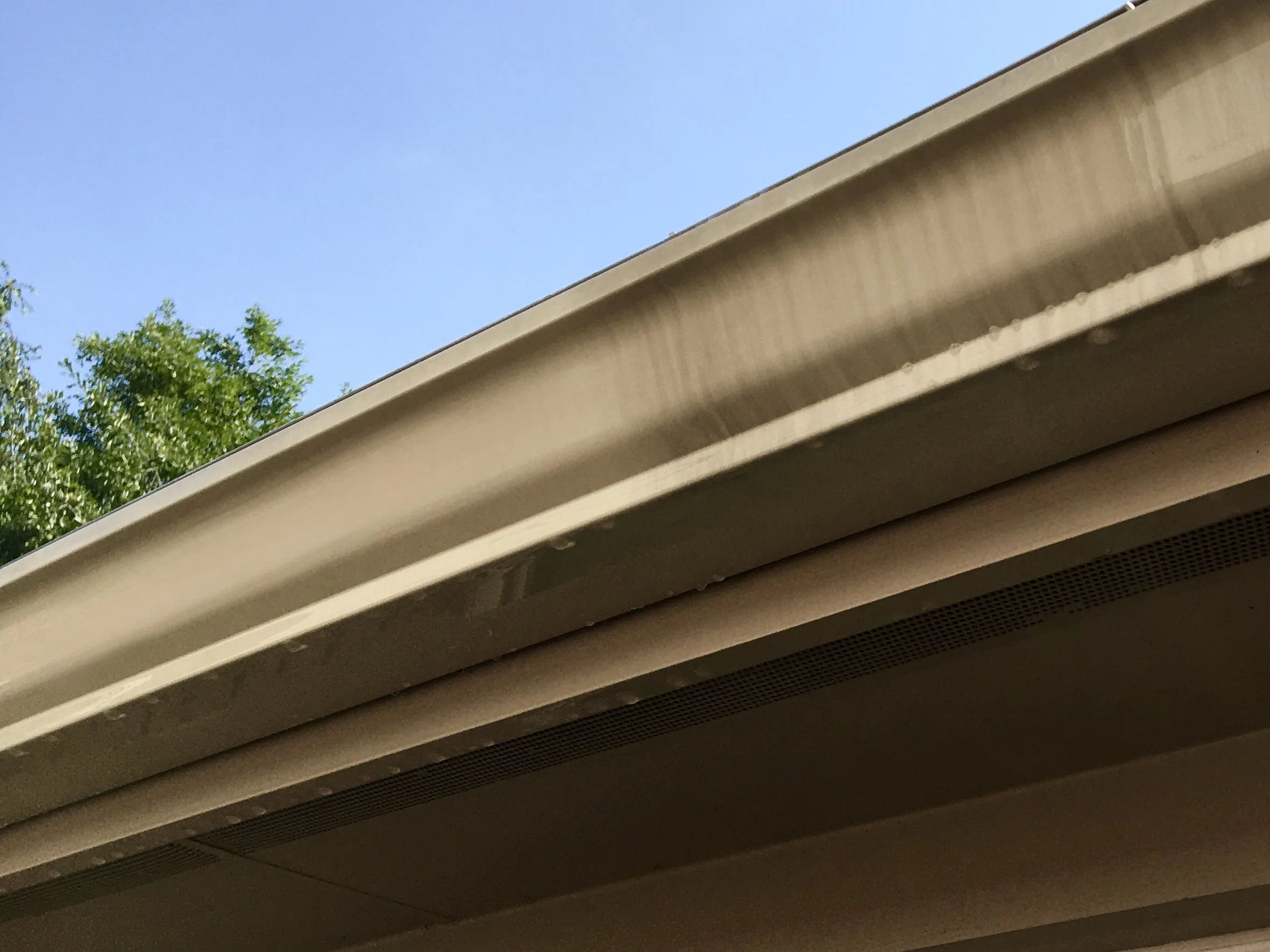 Close-up view of the edge of a beige outdoor awning or roof with a clear blue sky and some green trees in the background.
