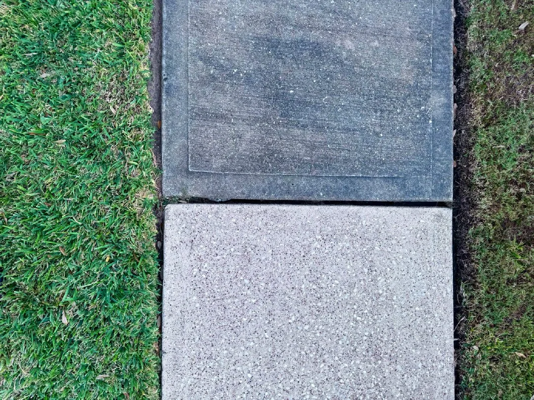 Close-up of two concrete sidewalk slabs, one gray and textured, the other darker and smoother, with grass on each side.