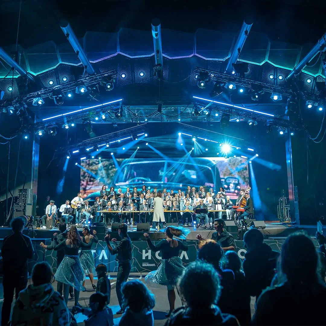 Image shows an outdoor stage lit with concert lighting as an audience watches a performance.