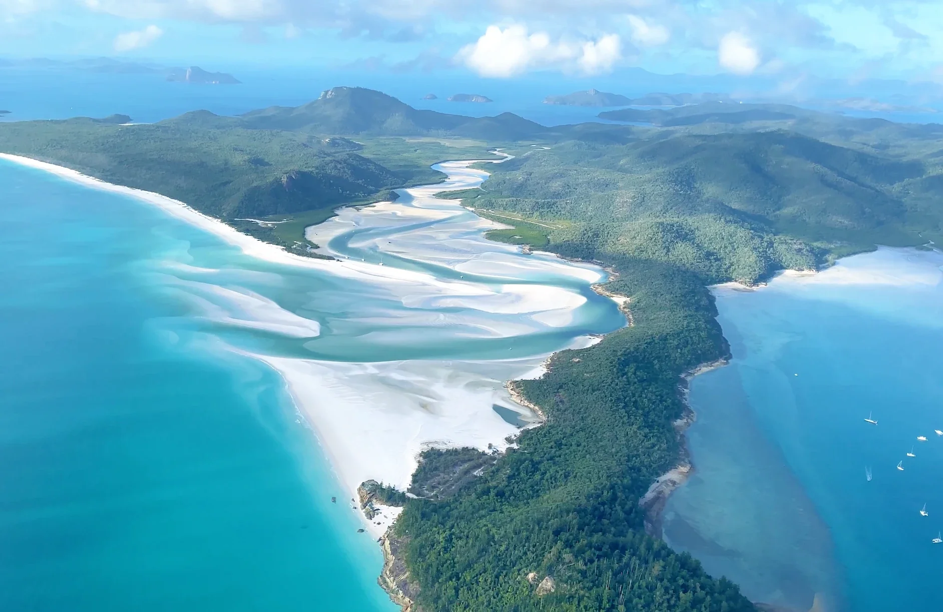 Aerial view of a coastal landscape with a winding river and white sand beaches, surrounded by lush green hills and distant islands under a partly cloudy sky.