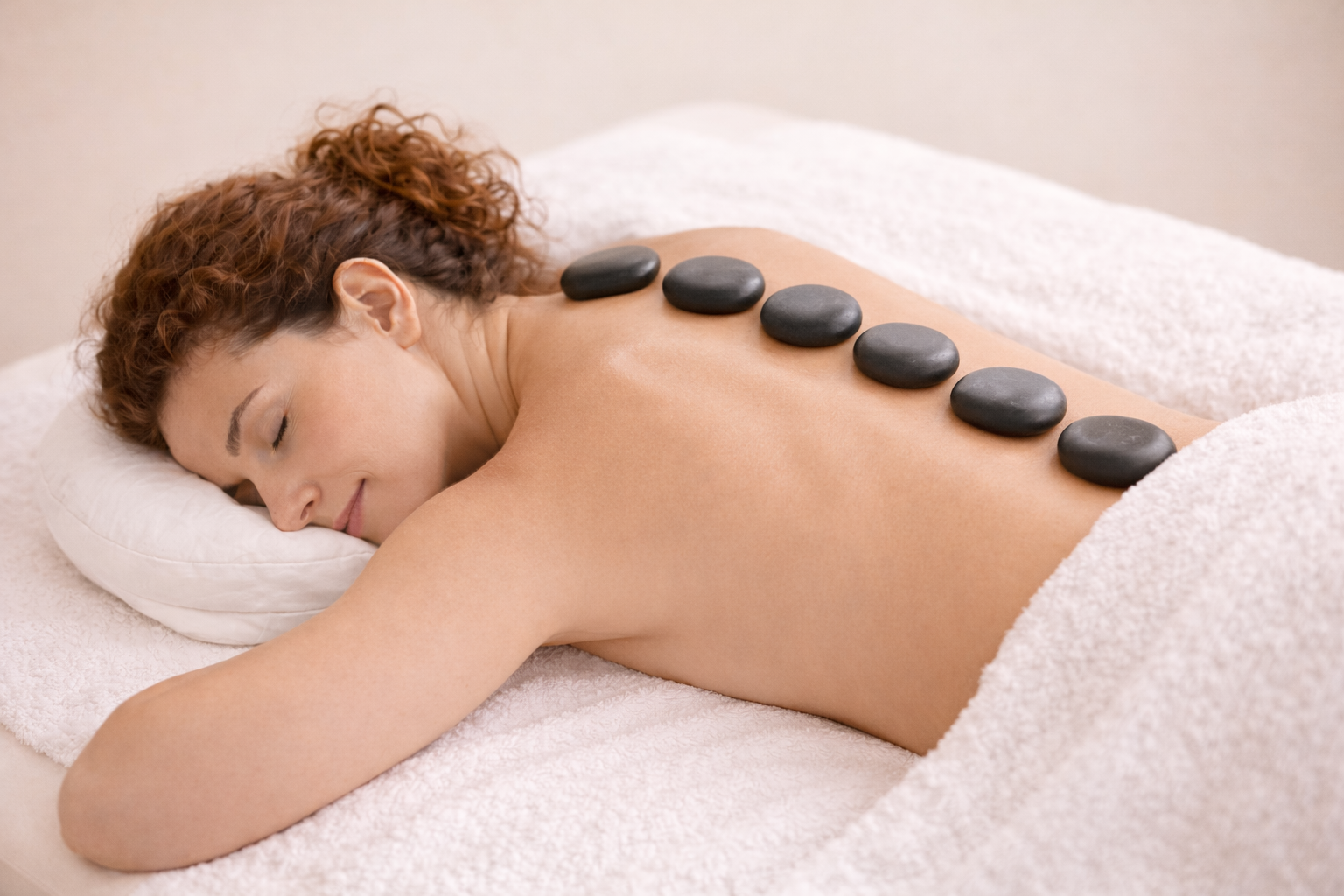 A woman with curly hair is lying face down on a massage table, resting her head on a pillow. Hot stones are placed along her back for a hot stone massage.