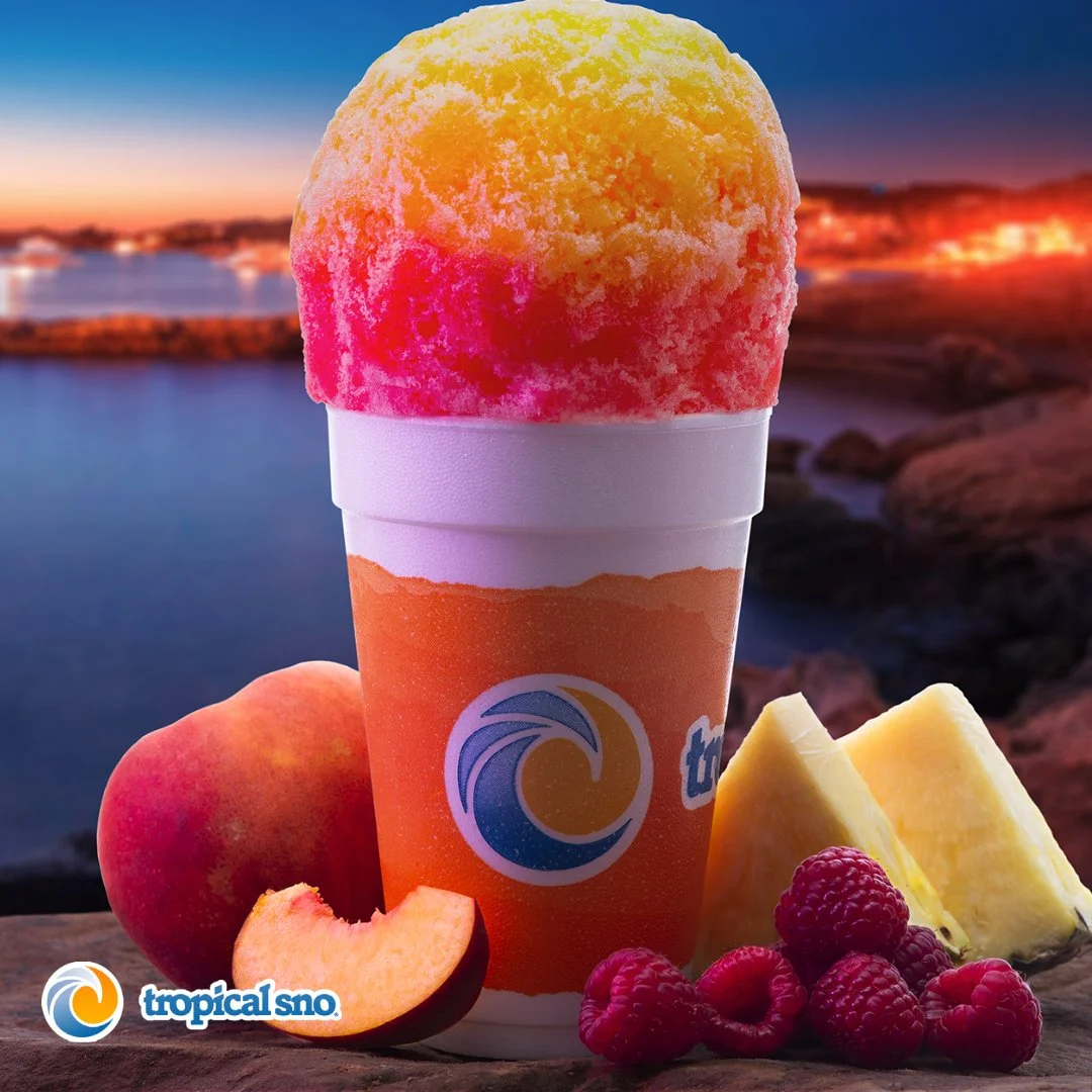 A tropical sno cone with yellow, pink, and red flavors in a cup held up against a sunset background over water, with peaches, pineapple, and raspberries in the foreground.