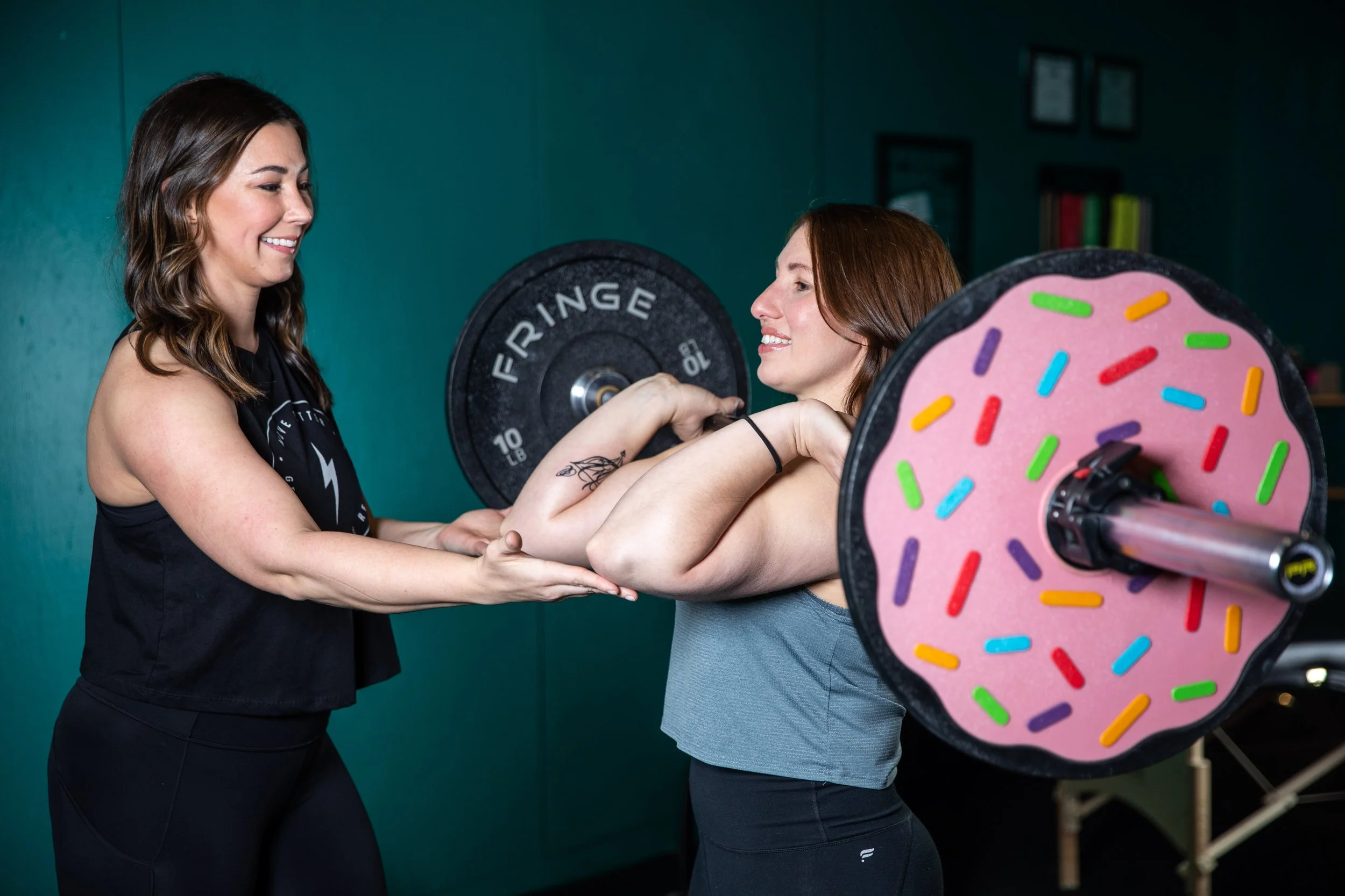 Two women in workout attire lifting a barbell with a pink donut-shaped weight with colorful sprinkles, while a trainer assists and encourages them in a gym environment.