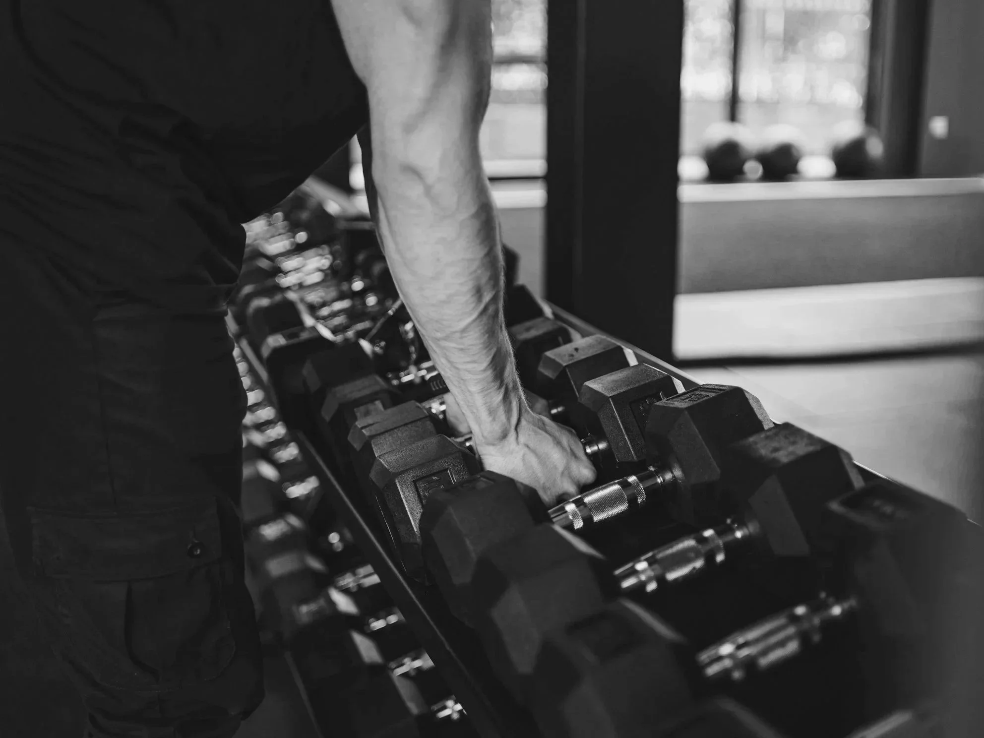 A person lifting a dumbbell in a gym, with a row of dumbbells on a rack beside them, in black and white photo.