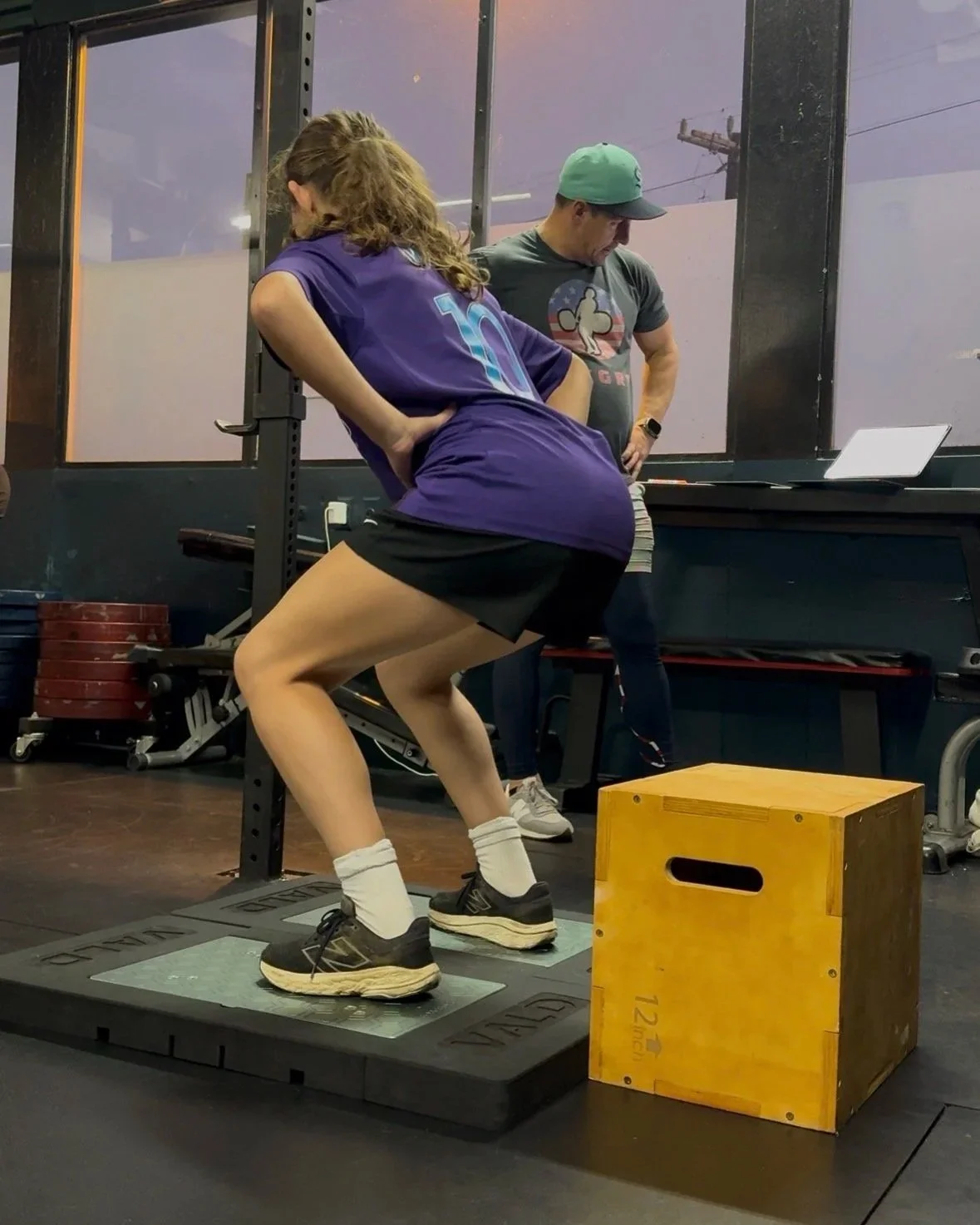 A woman in a purple sports jersey and black shorts is preparing to lift weights on a platform with her hands on her hips, while a man in a baseball cap and gray T-shirt looks on in a gym or workout space.