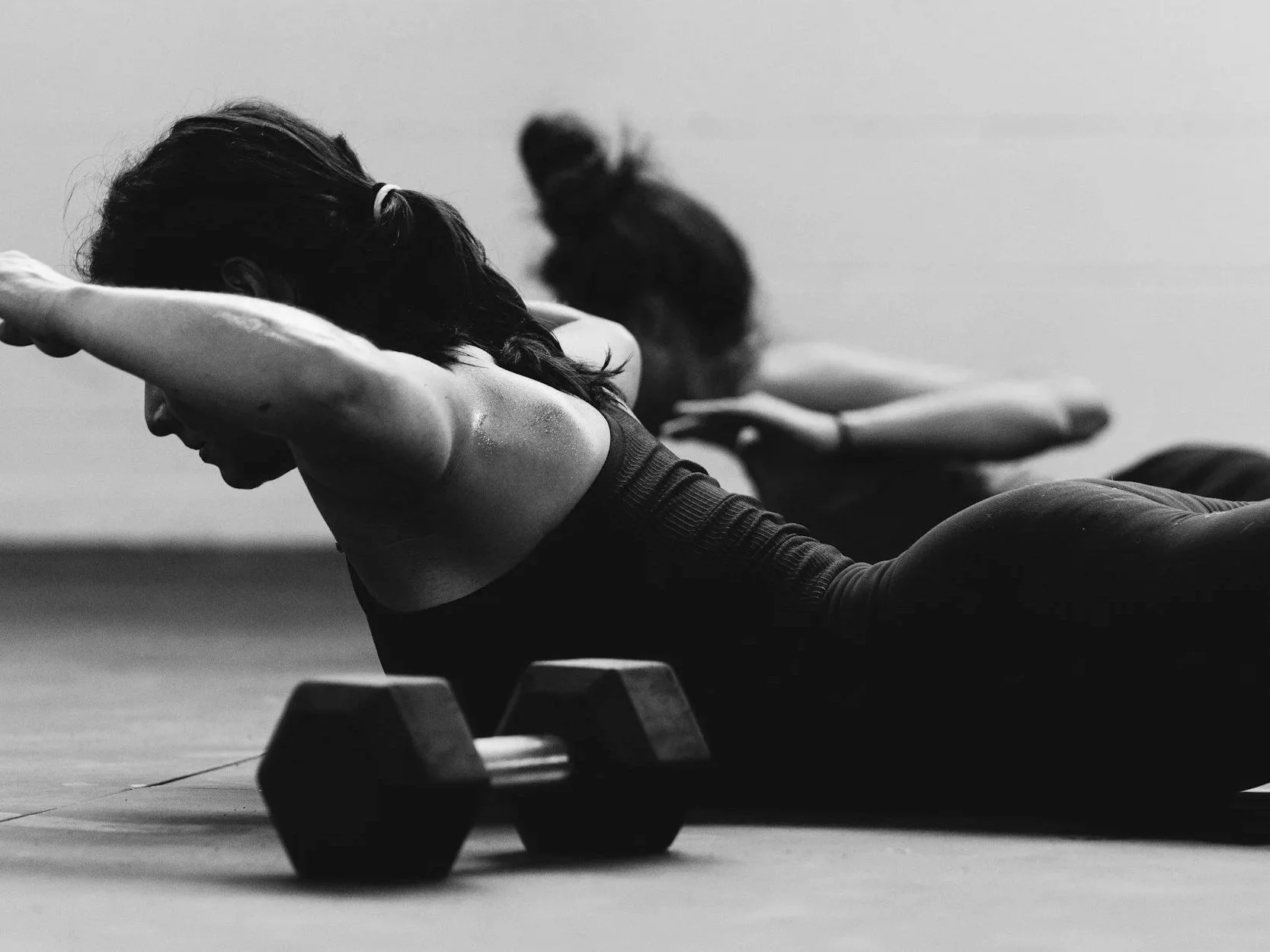 Two women performing a plank exercise with dumbbells on a gym floor in black and white.