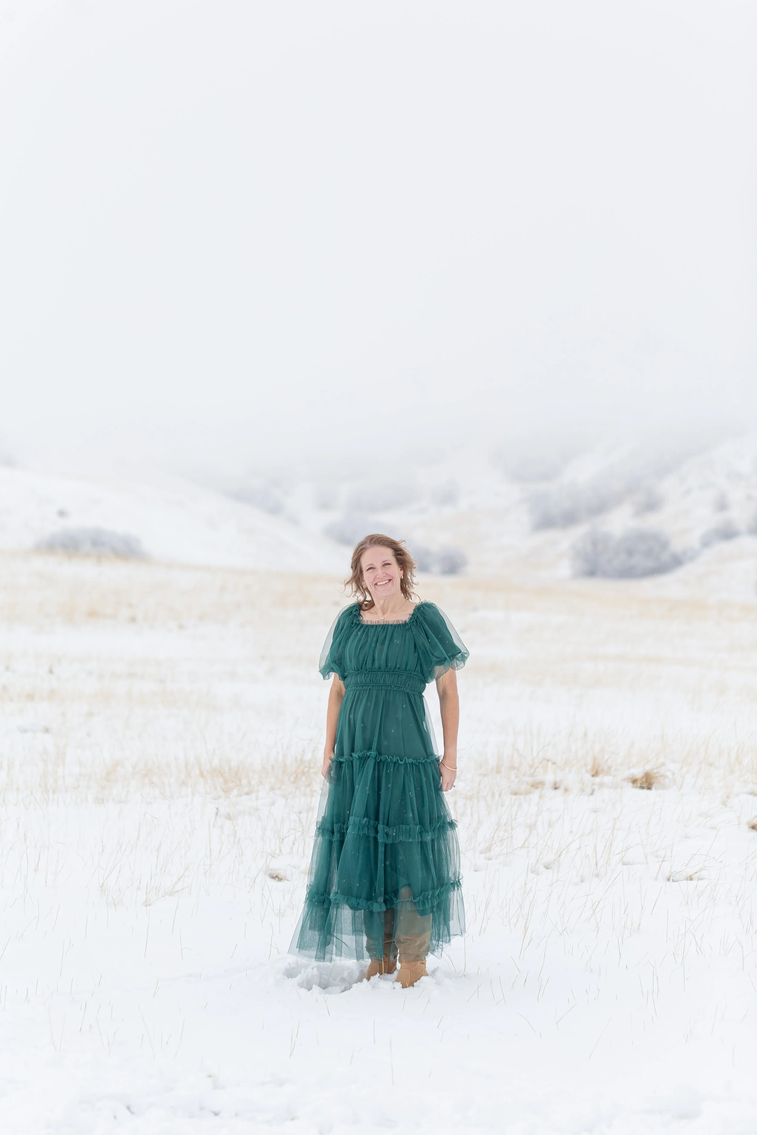 A woman in a long, green dress standing in a snow-covered field with a misty background.