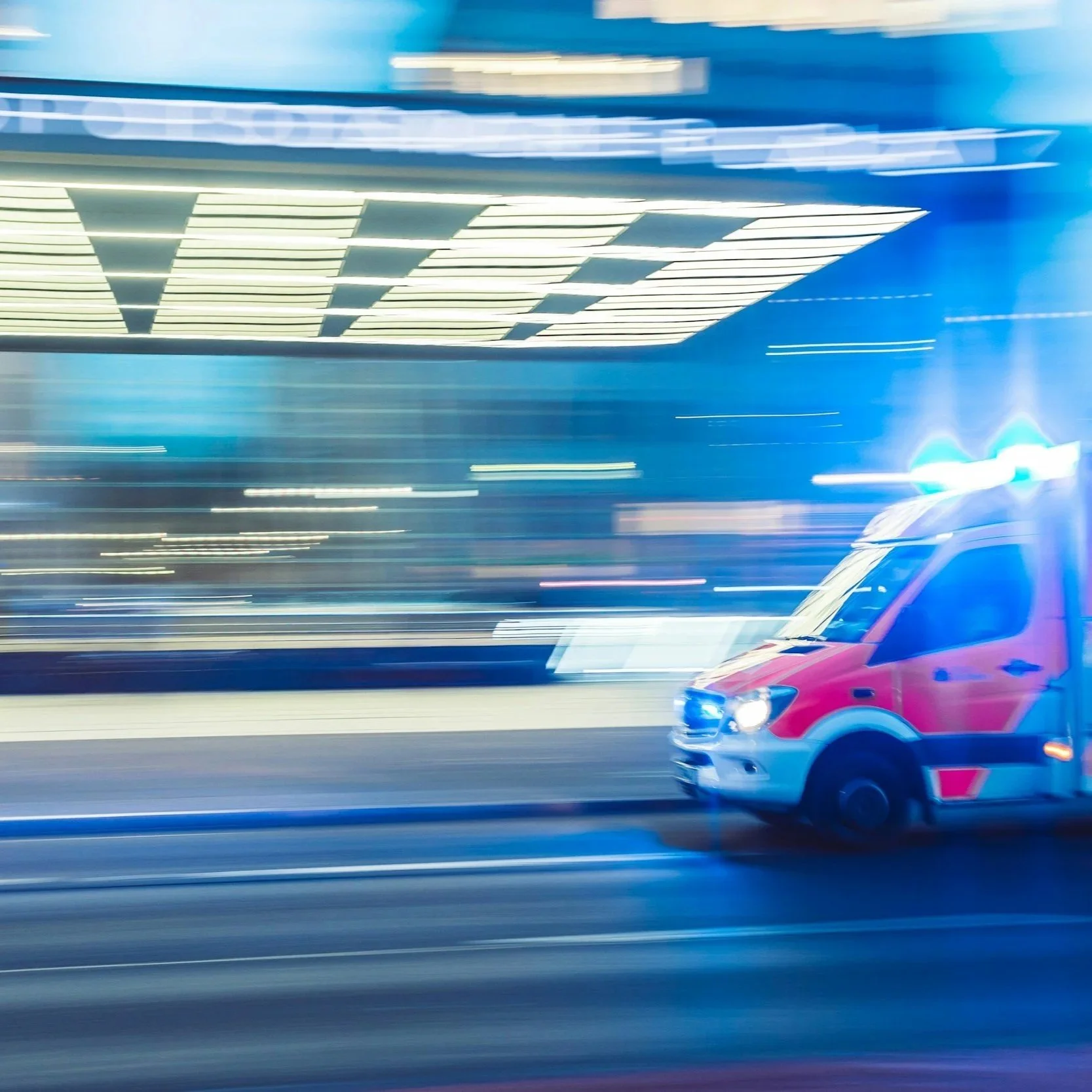 Blurred image of an ambulance driving at night, with flashing blue lights, in a city with illuminated buildings.