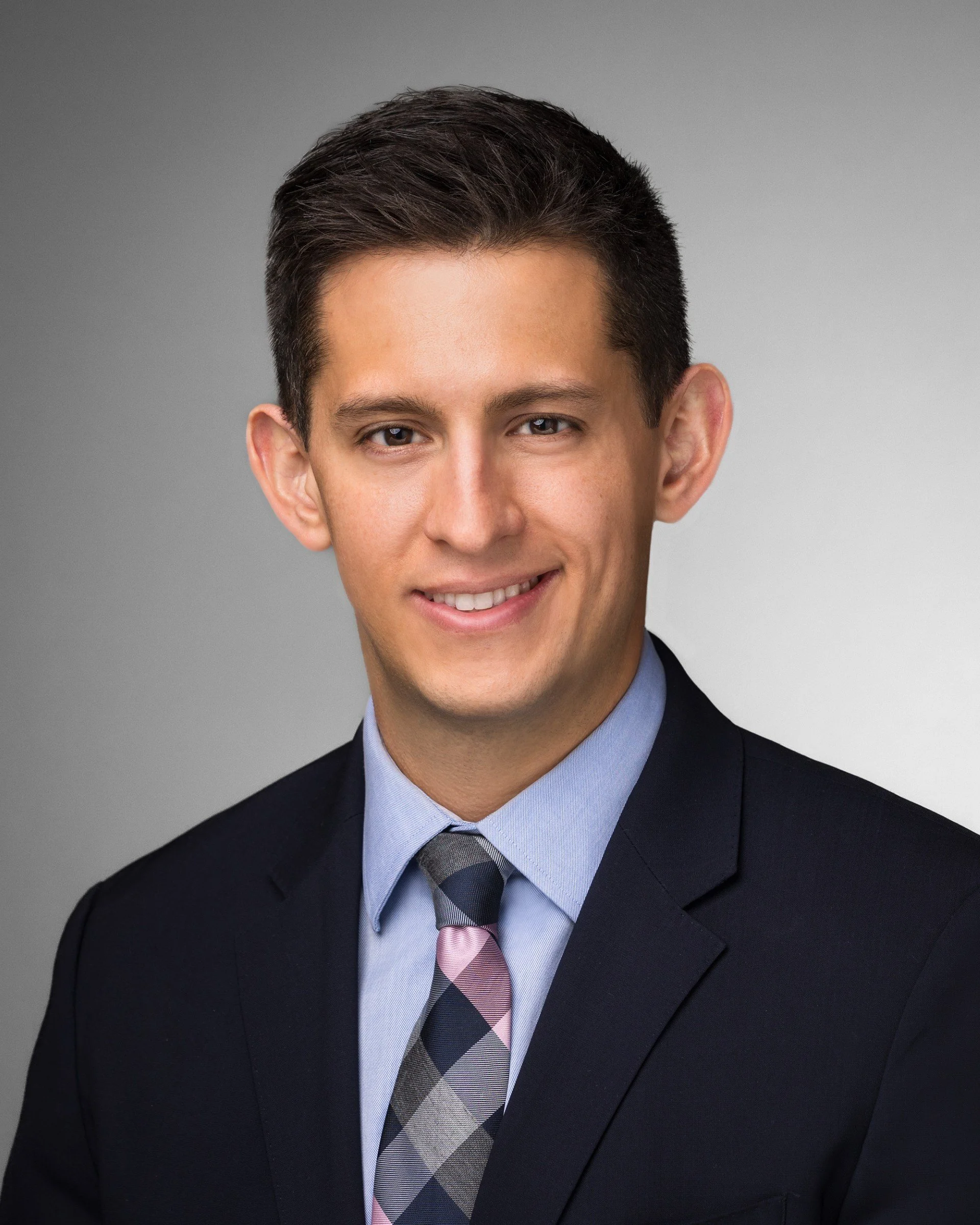 Professional headshot of a young man in a dark suit, light blue shirt, and a plaid tie, smiling against a gray background.