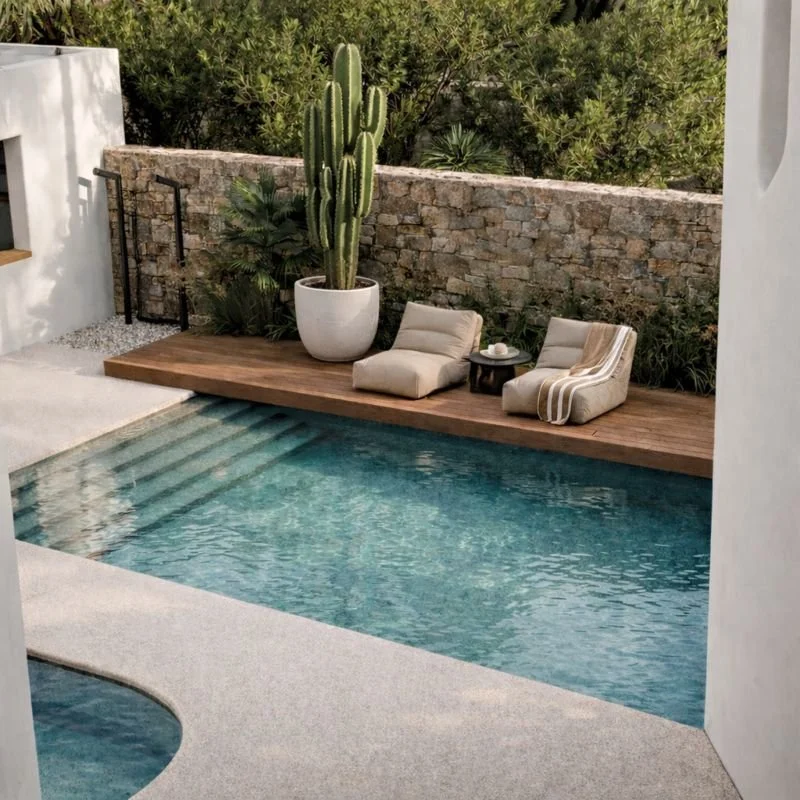 A stylish swimming pool with clear water, surrounded by a concrete deck with a wooden lounge area featuring cushioned chairs, a large potted cactus, and lush greenery in the background.