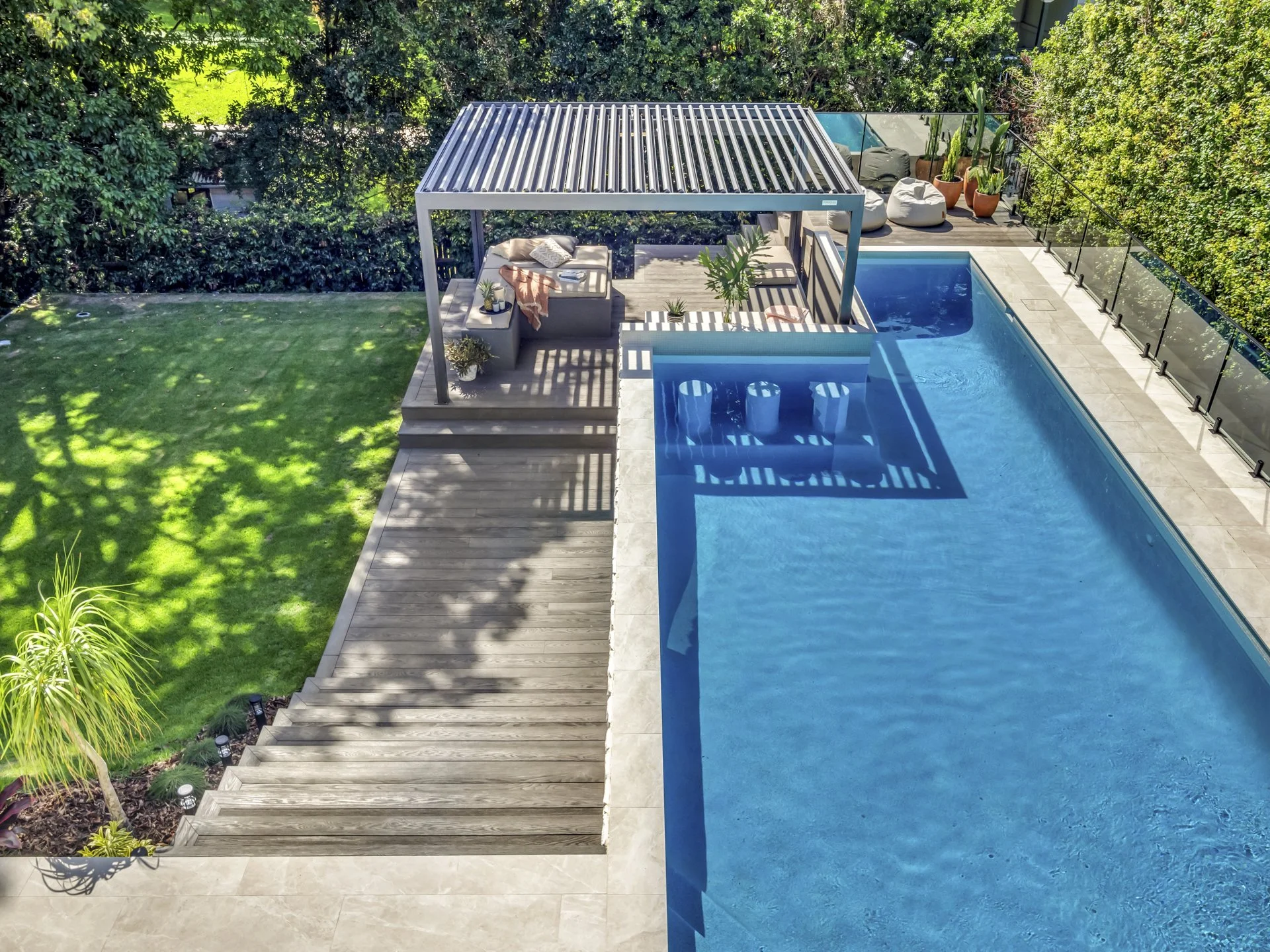 A backyard with a swimming pool, a wooden deck, and a shaded seating area with outdoor furniture, potted plants, and a glass fence.