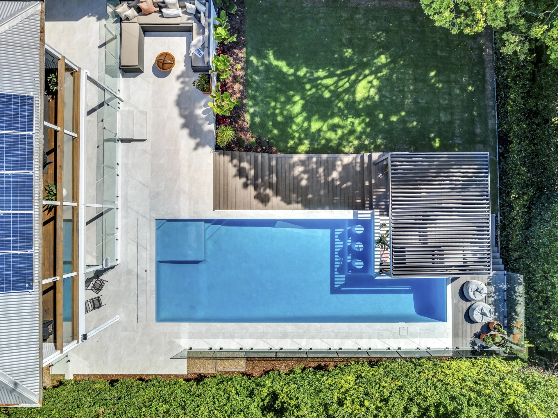 Aerial view of a backyard patio with a swimming pool, outdoor seating area, wooden deck, green grass, and surrounding trees and bushes.