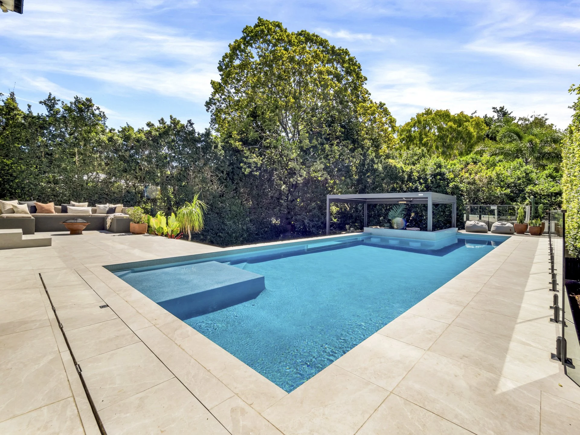 Modern backyard swimming pool with beige tiled deck, surrounded by lush green trees, outdoor seating with cushions on the left, potted plants, and a covered seating area on the right under a canopy.