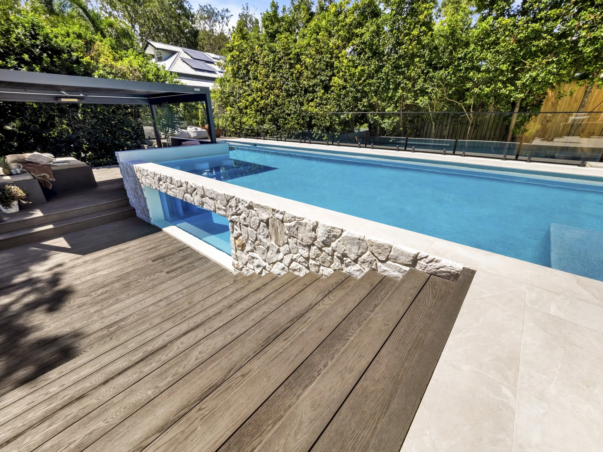 A modern backyard swimming pool with stone accents, surrounded by wooden decking and a tree-lined fence.