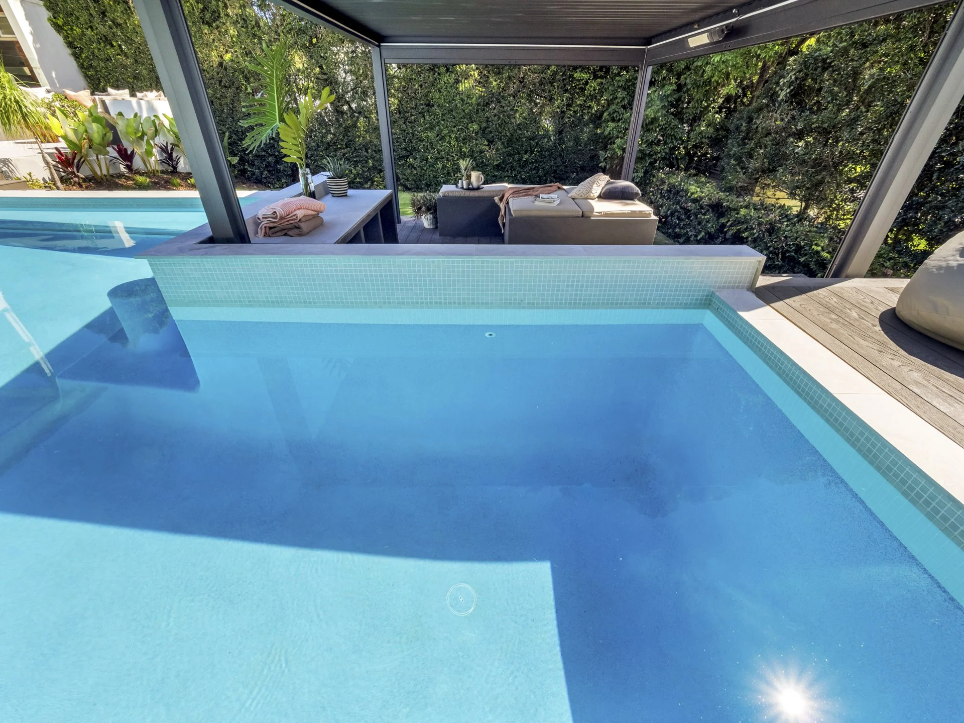 View of a small swimming pool with clear blue water, with a shaded outdoor seating area and lush green hedges in the background.