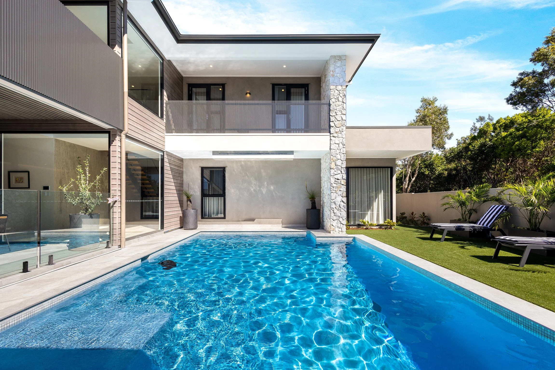 Modern two-story house with pool in backyard, outdoor lounge chairs, potted plants, and green lawn, under a blue sky.