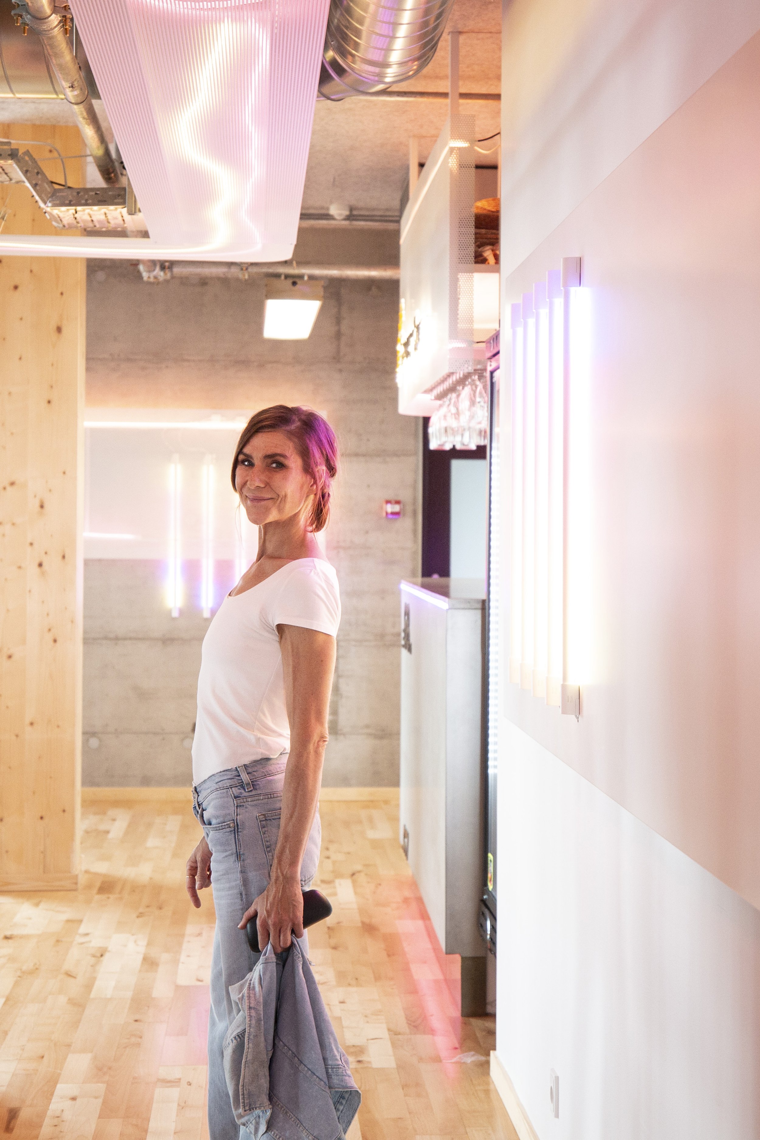 A woman with shoulder-length brown hair wearing a white t-shirt and light blue jeans, holding a denim jacket and a smartphone, standing in a modern, well-lit space with wooden flooring and pastel-colored LED lighting on the wall.