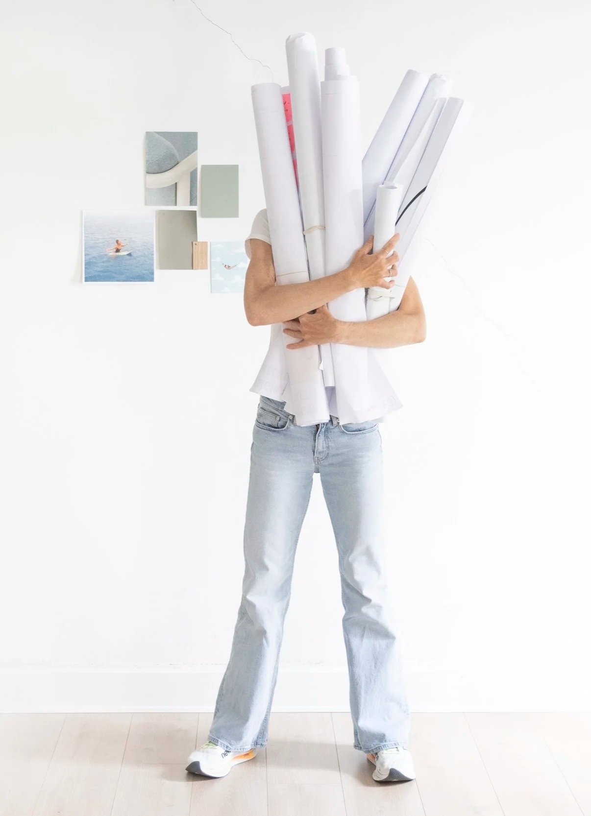 Person standing indoors holding a large bundle of rolled-up papers, partially obscuring their face. The individual is wearing light-colored jeans and sneakers. The background features a white wall with small decorative panels and photographs of water activities.