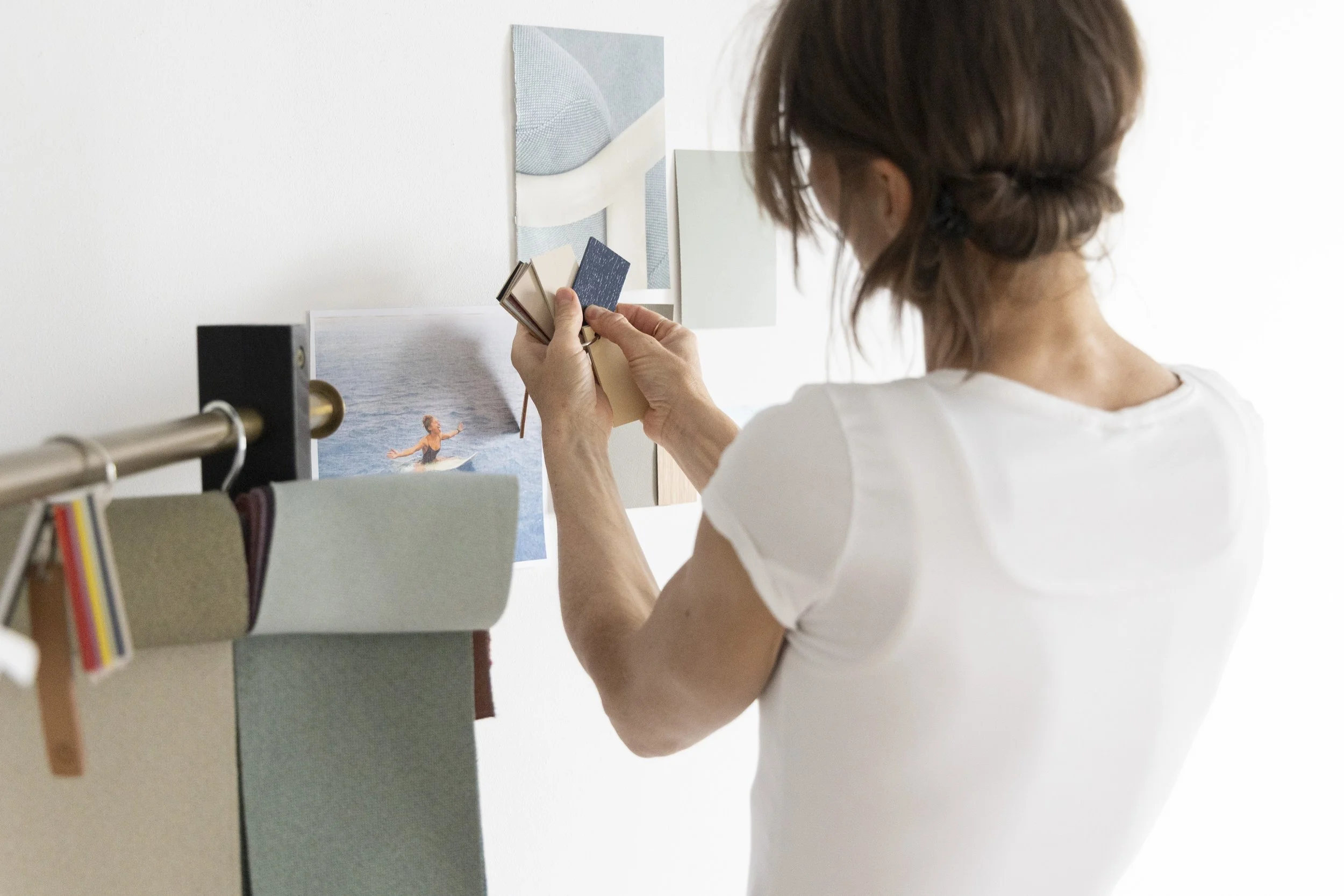 Woman with brown hair arranging fabric swatches on a wall above a photograph of a person surfing.
