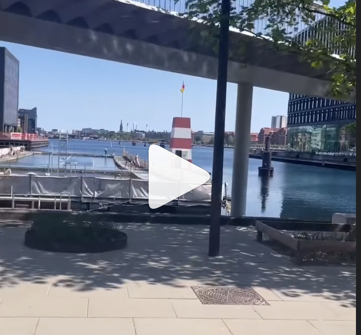 A waterfront harbor with a lighthouse, docked boats covered with white tarps, and a city skyline with tall buildings, under a clear blue sky.