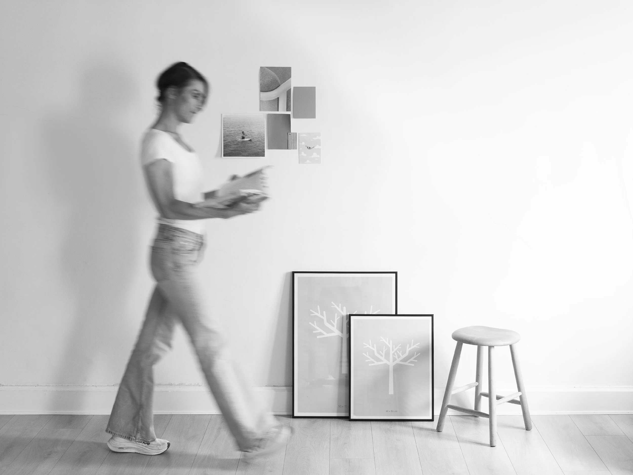 A woman walking in a gallery space, looking at art. Artworks and framed pictures are on the wall and floor, including paintings of trees and a photograph of a person on a boat. A wooden stool is nearby.