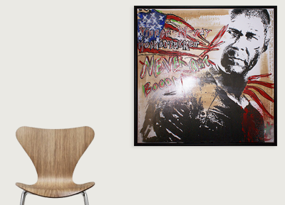 A framed poster of a man with a serious expression, street art style, with American flag motifs, words like 'BOOM!' and 'Never' in red and yellow, hung on a white wall next to a wooden chair.