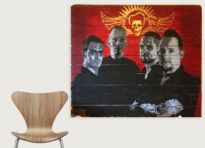 A wall painting of four men in black suits with a red background featuring a skull with wings and a halo. In front of the wall is a modern wooden chair.