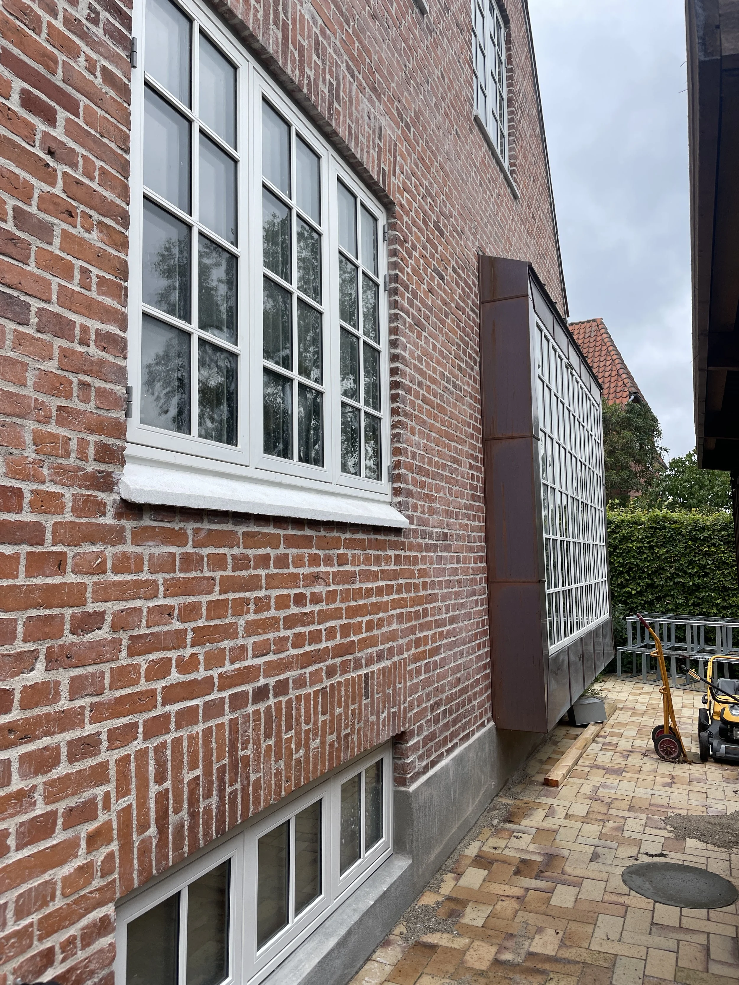 Udsigt af det ydre af en bygning med røde mursten, hvide vinduer og en udbygning med store glasruder, samt en stenbelagt terrasse med nogle haveværktøj og byggematerialer.