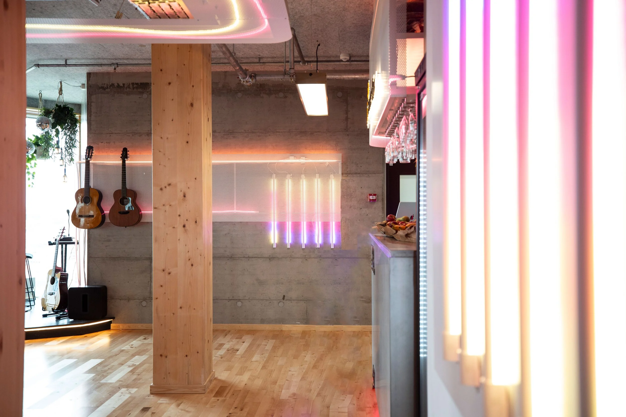 Indendørs musikbar med guitarer på væggen, moderne neonbelysning, træbjælker og et barområde med glas og snacks.