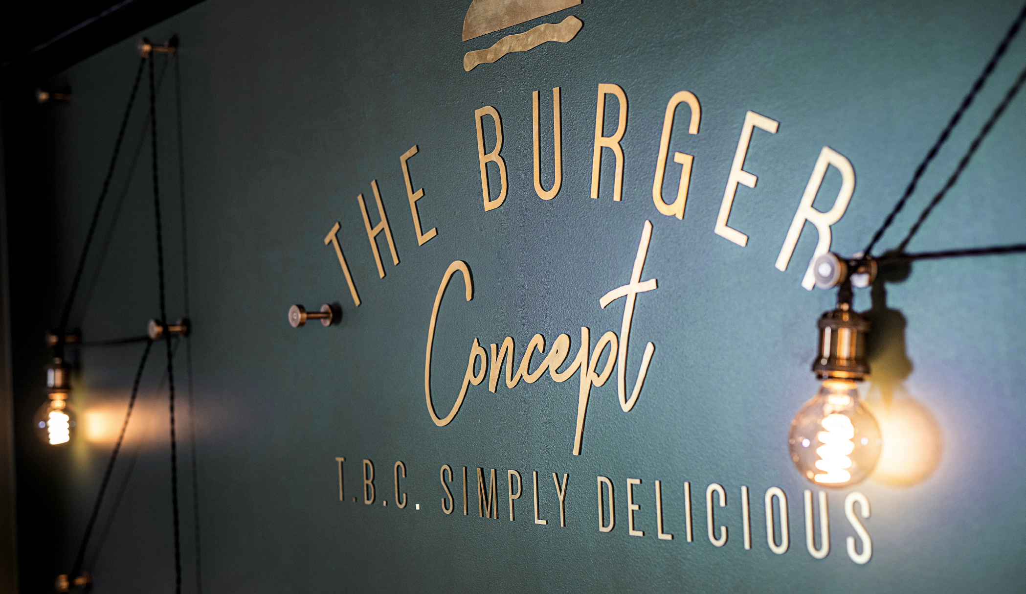 Vægdekoration med teksten 'The Burger Concept' og 'T.B.C. Simply Delicious' samt to lysende filament pærer, monteret på en mørkegrøn væg.