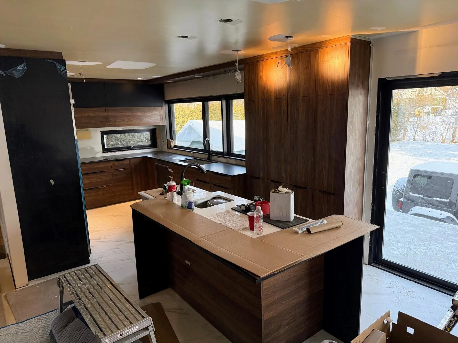 Modern kitchen under construction with wooden cabinets, black countertops, large windows, construction tools, and materials.