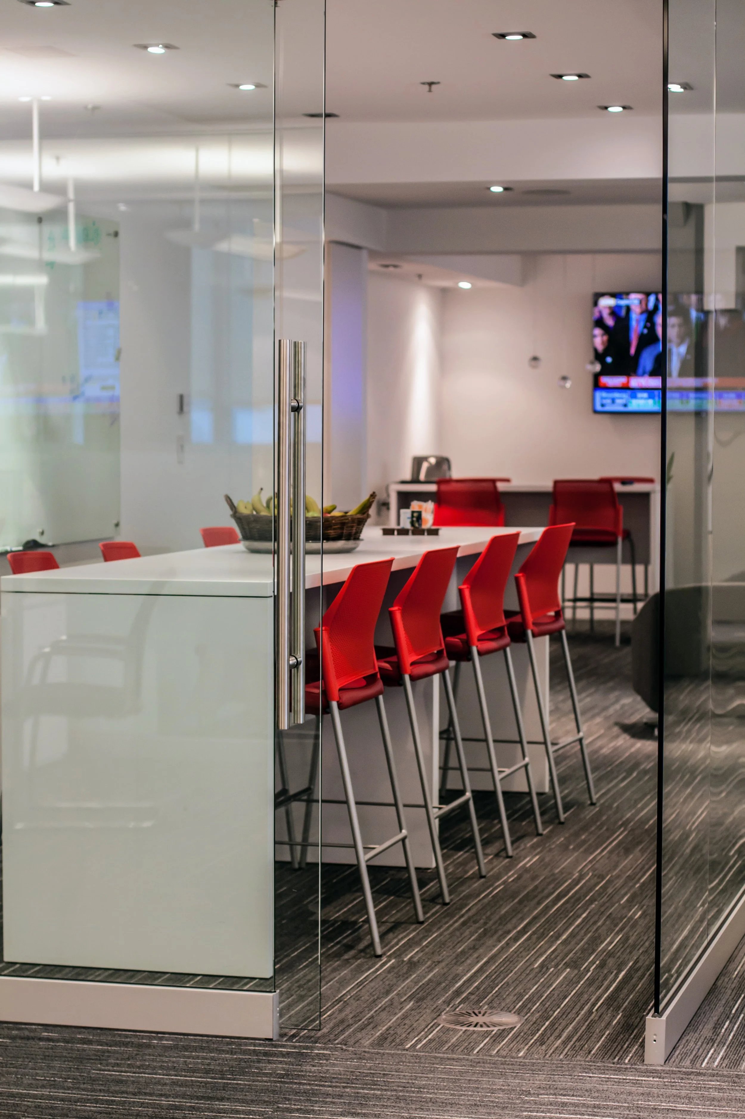 Empty conference room with white table, red chairs, and a basket of bananas, enclosed by glass partitions, with a TV screen on the wall showing news.