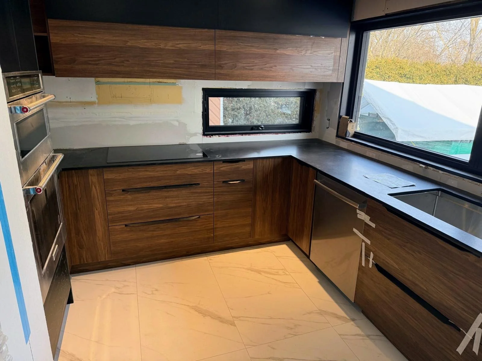 Modern kitchen under renovation with dark wood cabinets, black countertops, a sink, and two large windows providing natural light.
