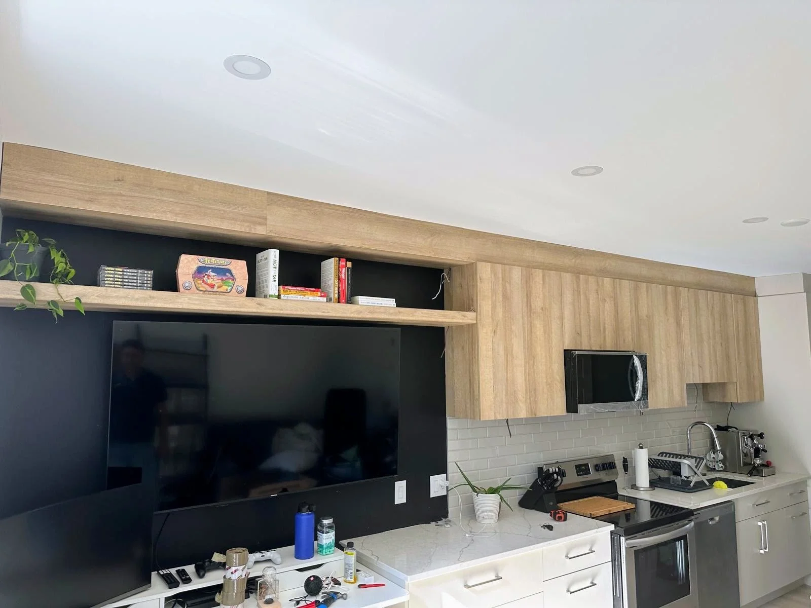 Modern kitchen with white cabinets, black backsplash, wooden upper cabinets, and a marble countertop. Items on the counter include a potted plant, paper towel, dish rack, soap dispenser, and various kitchen utensils. A large flat-screen TV is mounted