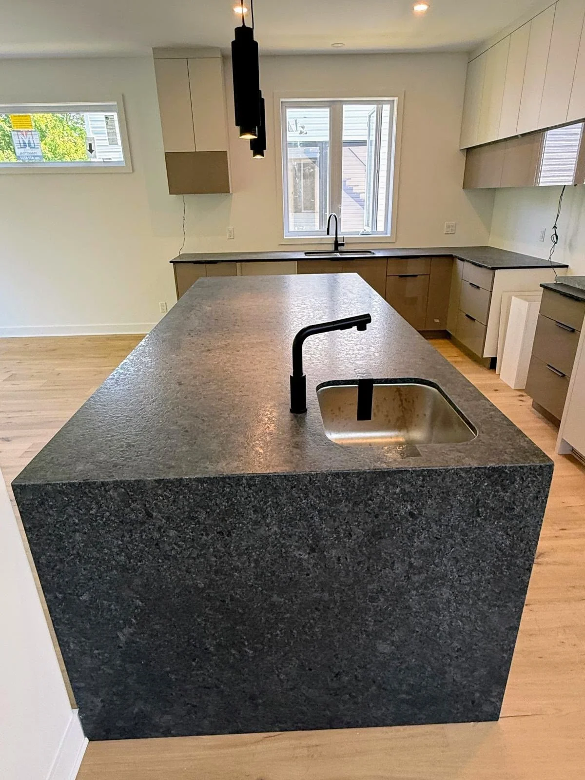Modern kitchen with black stone countertop island, black sink, black faucet, light wood cabinetry, two black pendant lights, and a window above the sink in a bright room.