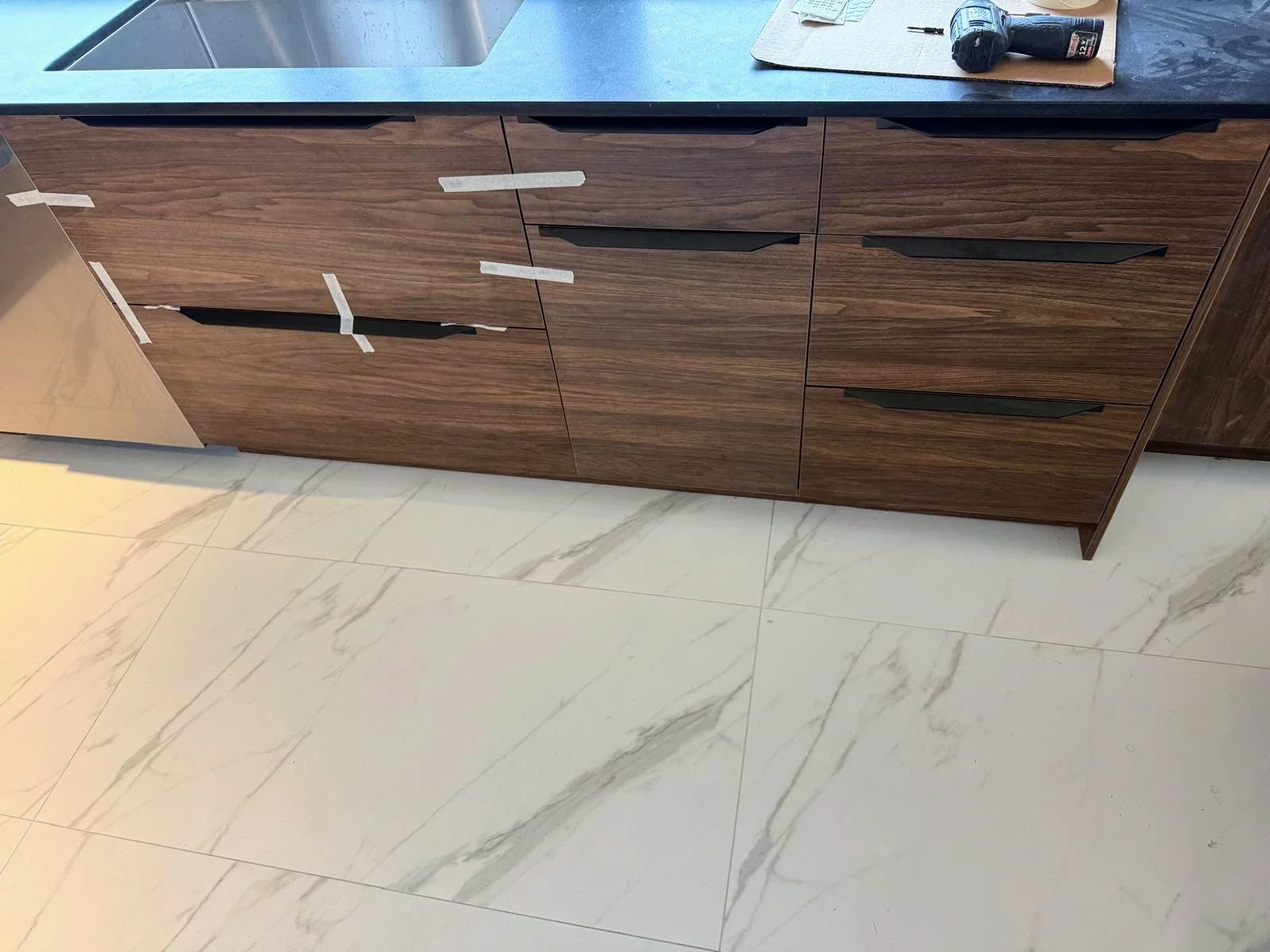 A kitchen countertop with a sink, counter space, and wooden cabinets with black handles, some of which are taped. A power drill and a notebook are on the countertop.