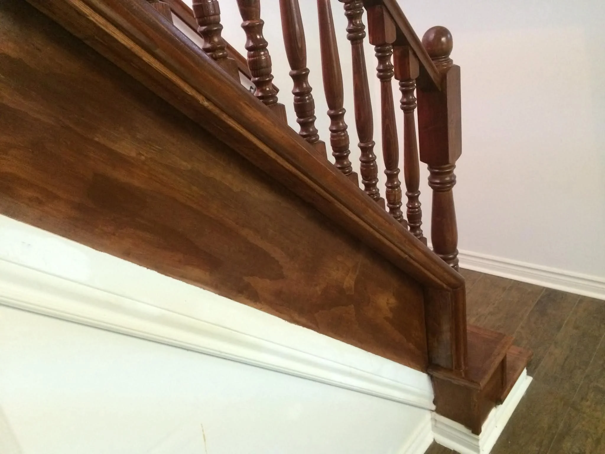 Close-up of a wooden staircase railing with turned spindles and a wooden handrail, next to a white wall and baseboard.