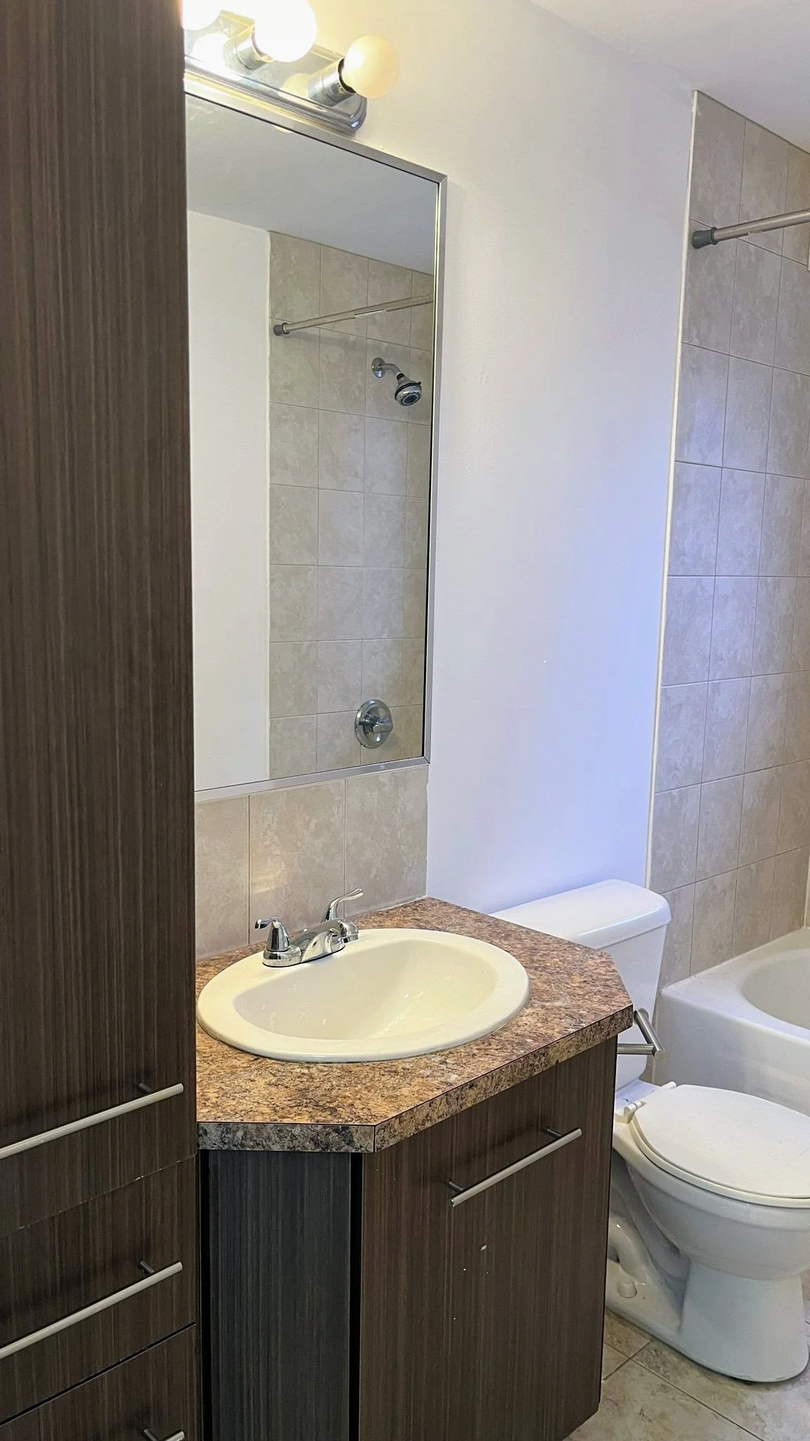 A bathroom with a dark wood vanity, a beige countertop, a white sink, a mirror above, and a partially visible toilet and bathtub. The shower has beige tile walls and a curtain rod. The lighting fixture has three bulbs.