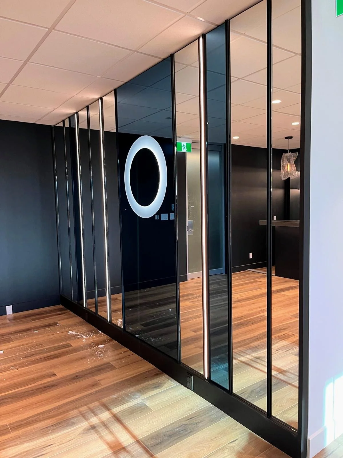 Interior of a modern office or commercial space with black walls, wooden flooring, and glass partition walls with metal frames. There is an illuminated circular sign on the wall and a green exit sign above a door.