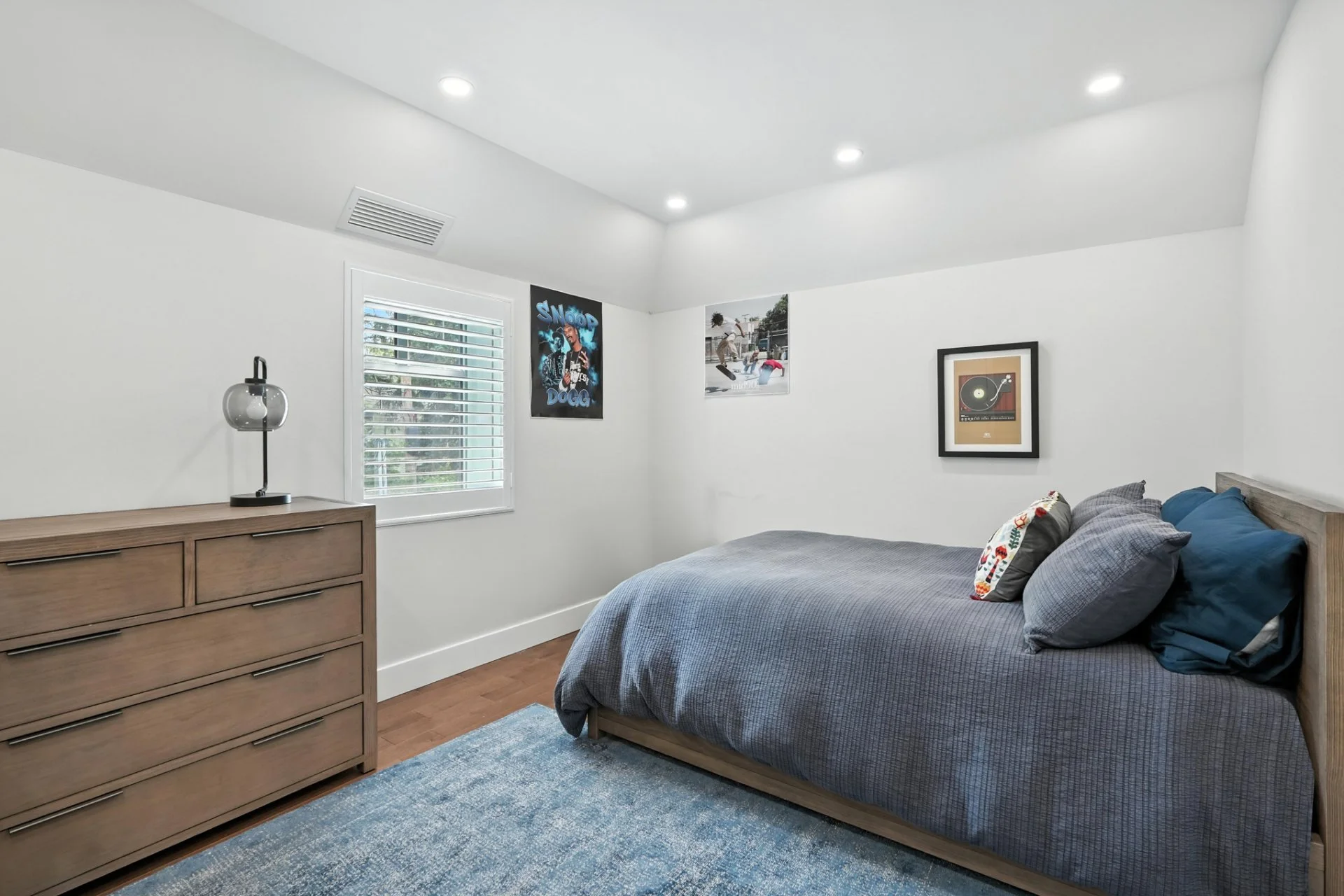 A bedroom with white walls, a wooden dresser, a bed with gray bedding, a window with white shutters, and music-themed wall art.