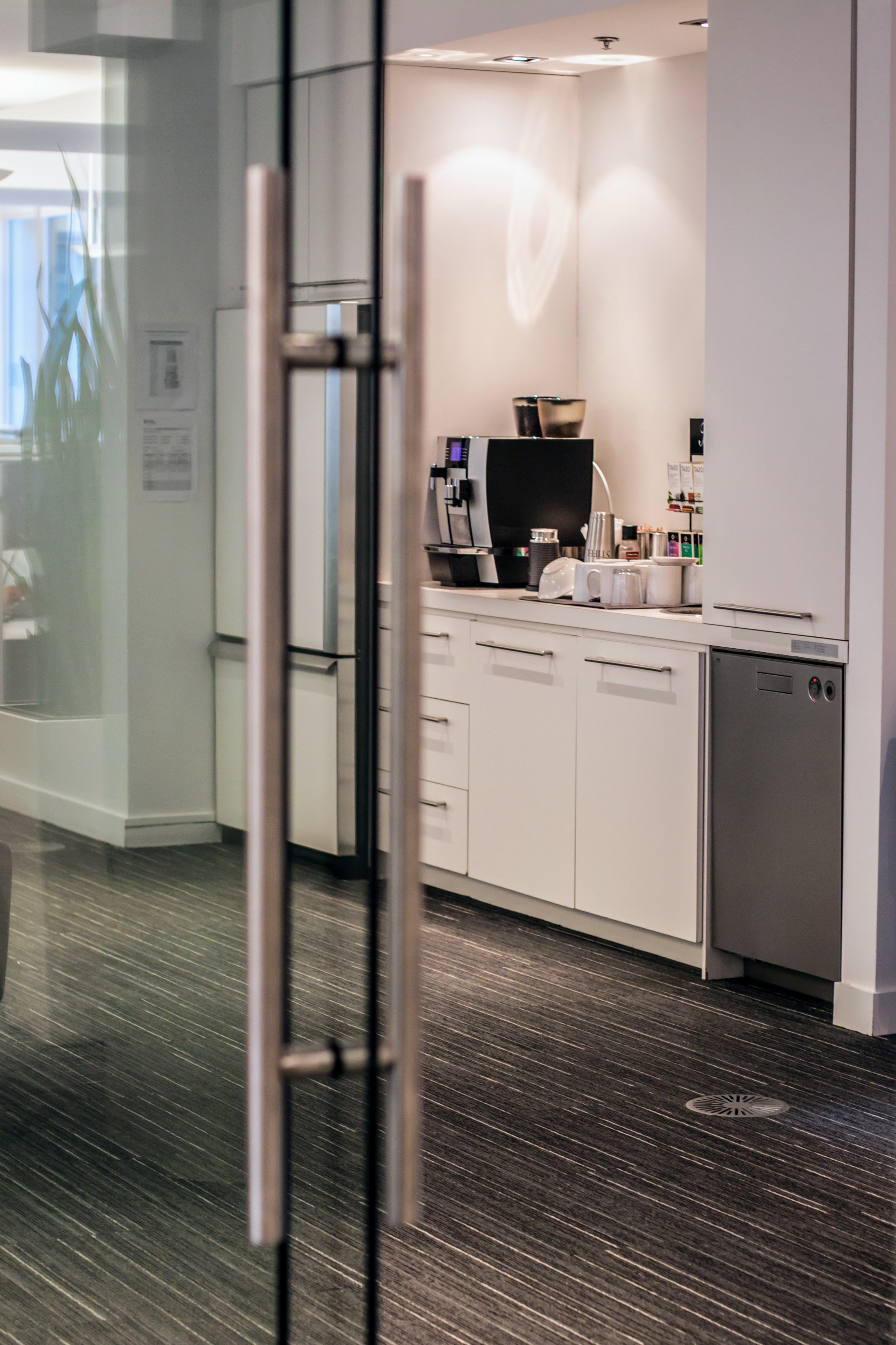 View of a coffee station in an office or break room with a coffee machine, cups, and coffee supplies on white cabinets, seen through a glass door with metal handles.