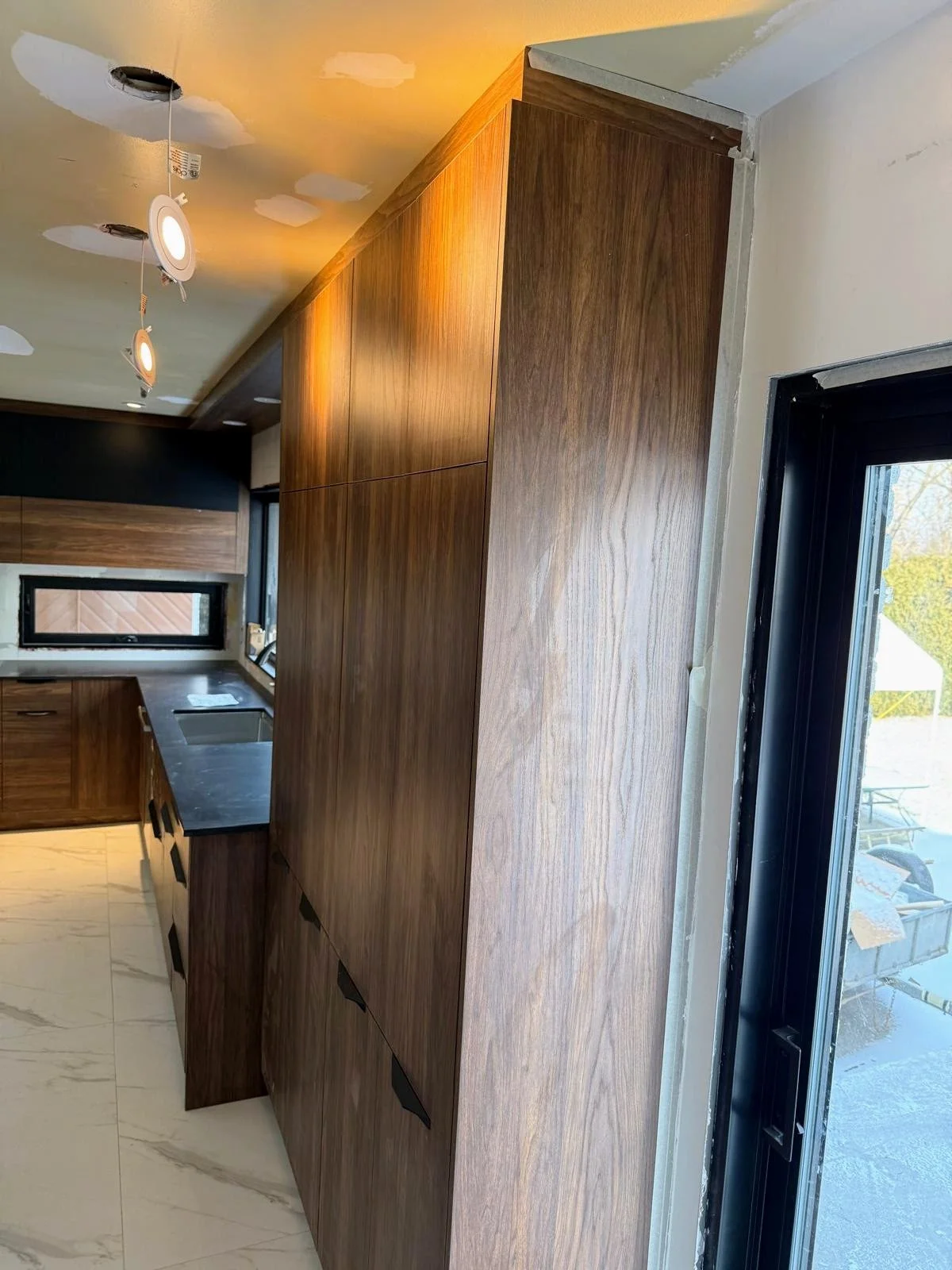 Interior view of a kitchen with wooden cabinets, a black countertop, a window, and a sliding glass door. The ceiling has track lighting, and some areas are unfinished.
