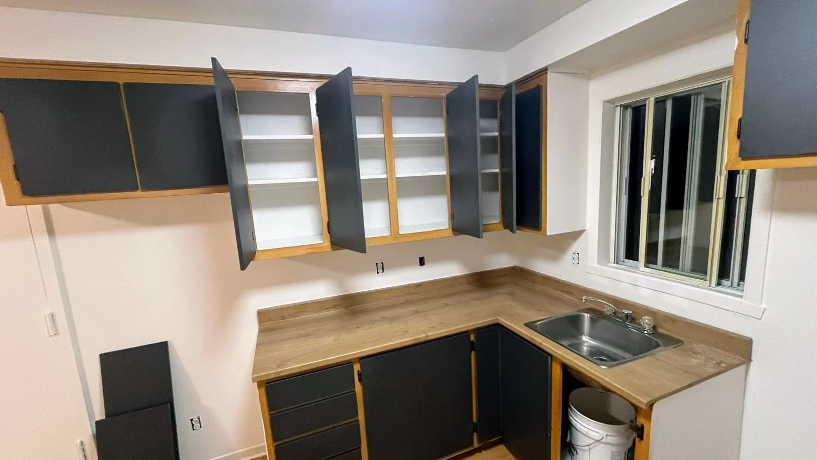 Empty kitchen with wooden cabinets, some open and some closed, a stainless steel sink under a window, and a white bucket near the cabinets.